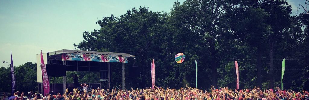 Color Run 5K massive crowd with arms raised toward an Apex stage under summer trees