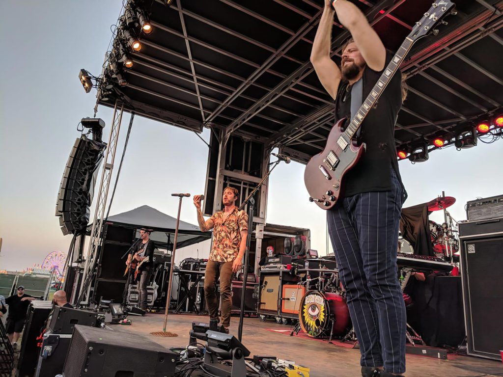 Band performing on Apex stage at golden hour county fair with ferris wheel in background