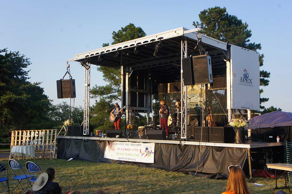 APEX 2016 mobile stage at an outdoor benefit concert, with a duo performing and Apex Stages branding on the side panel
