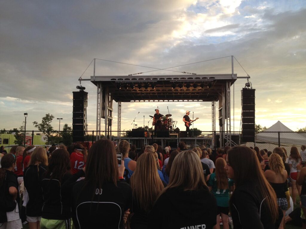 Crowd watching a band on the APEX 2420 mobile stage at sunset, with line array speakers flown on both sides