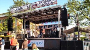 APEX 2420 mobile stage set up on a downtown street for a live music event, with sponsor banners on the roof