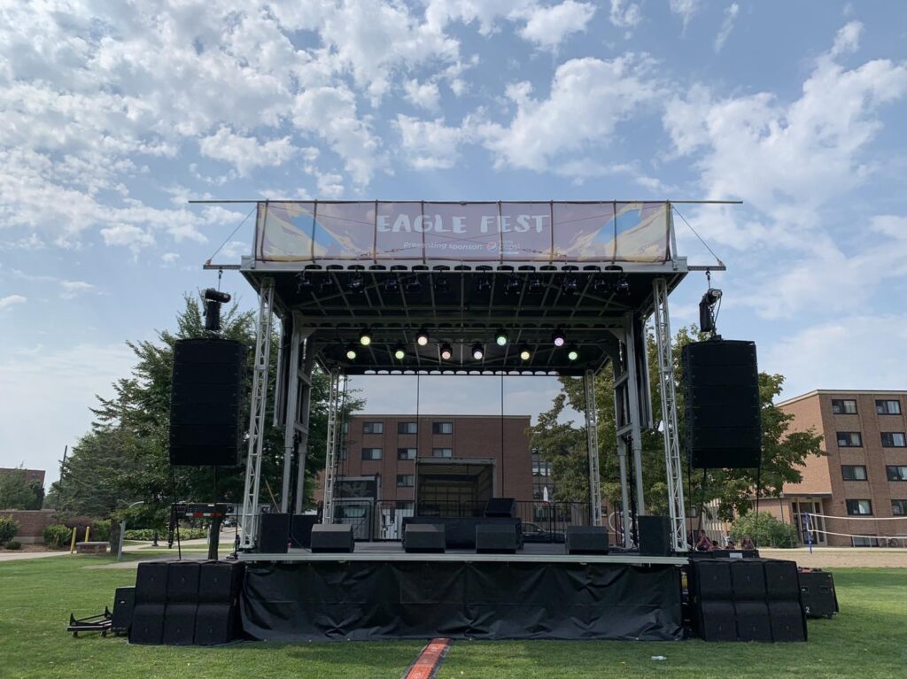 Front view of the APEX 2424 mobile stage on a lawn, with an Eagle Fest banner on the roof header and speakers stacked at deck level