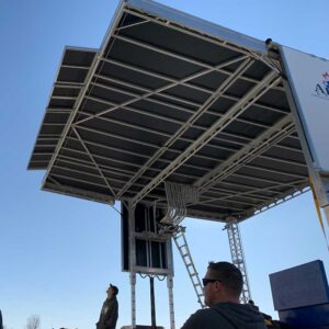 APEX 2424 mobile stage roof canopy raised into position against a clear sky as crew look on